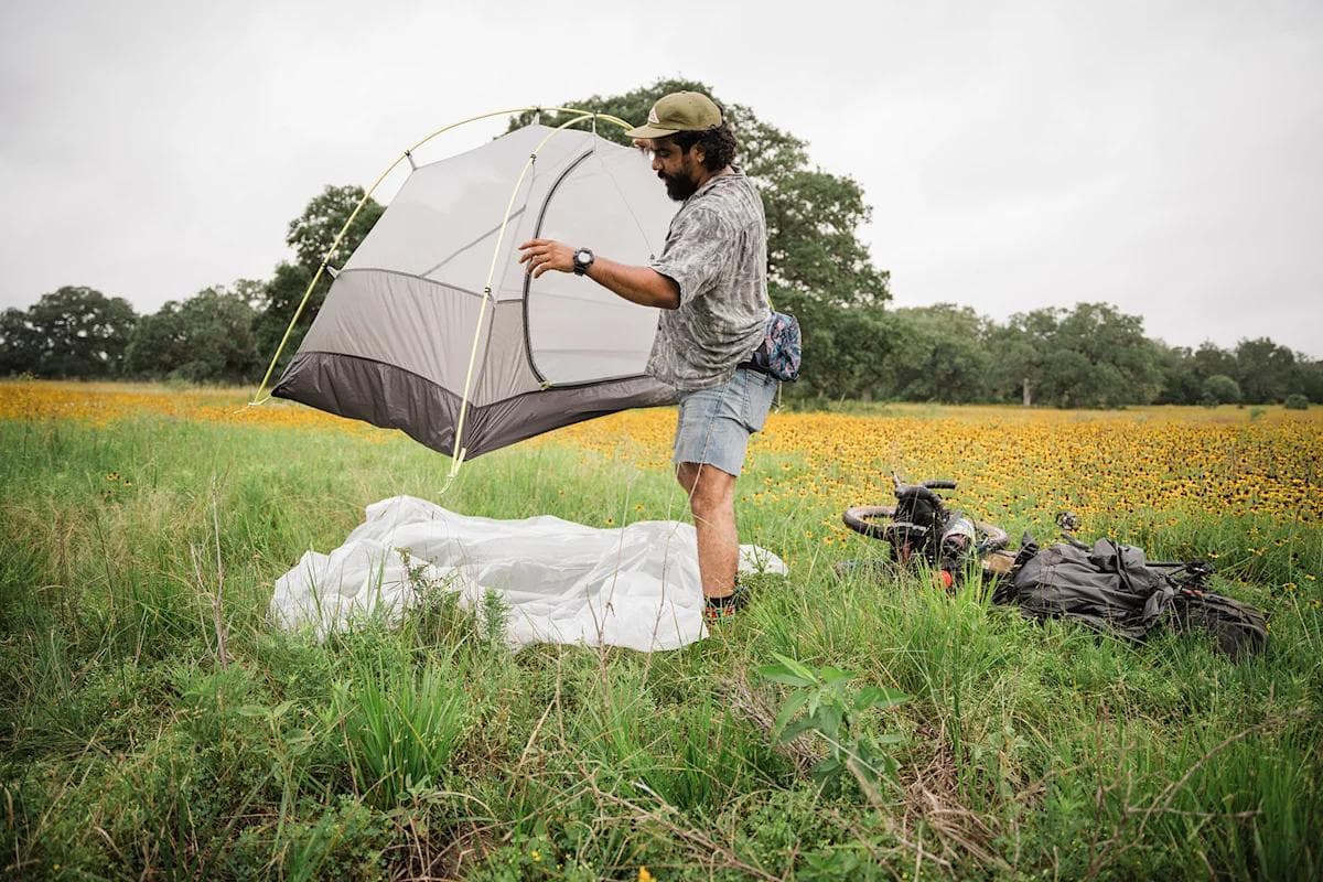 A man sets up a gray tent in a field of yellow wildflowers next to a bikepacking bicycle.