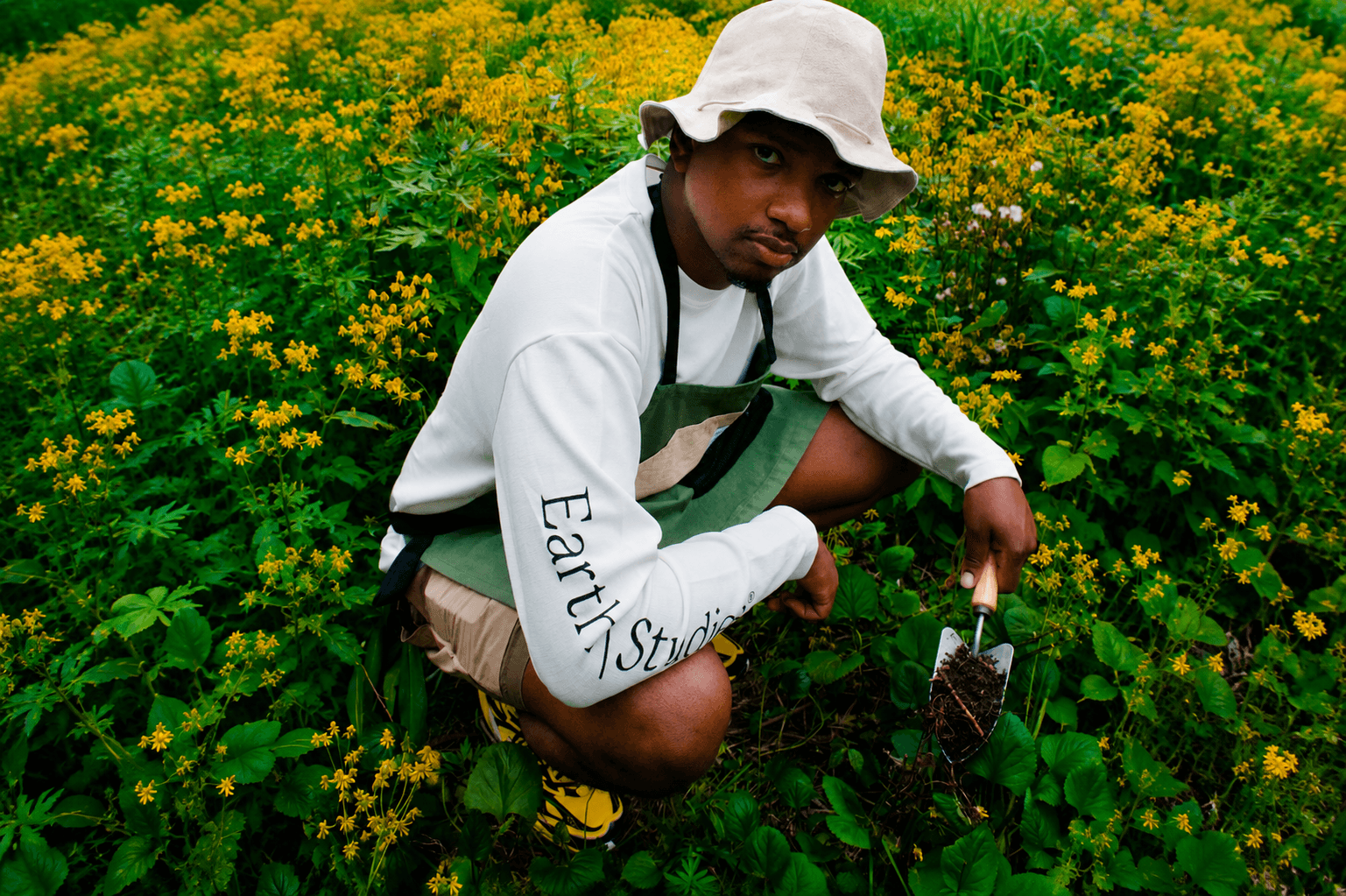 A man in a bucket hat, white "Earth Studios" shirt, and green apron squats in a flowery field, holding a trowel with soil.