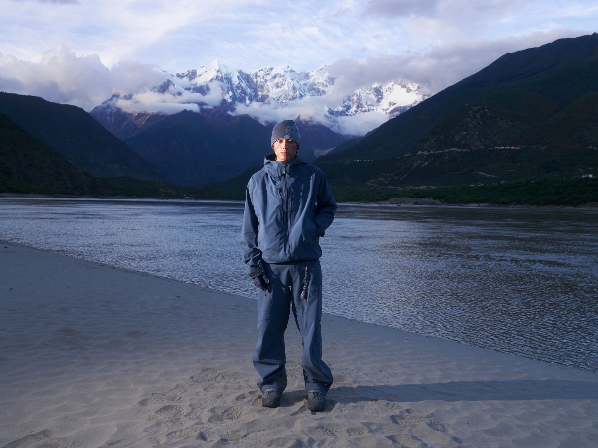 Man in blue winter gear on a sandy riverbank with snowy mountains and cloudy sky.
