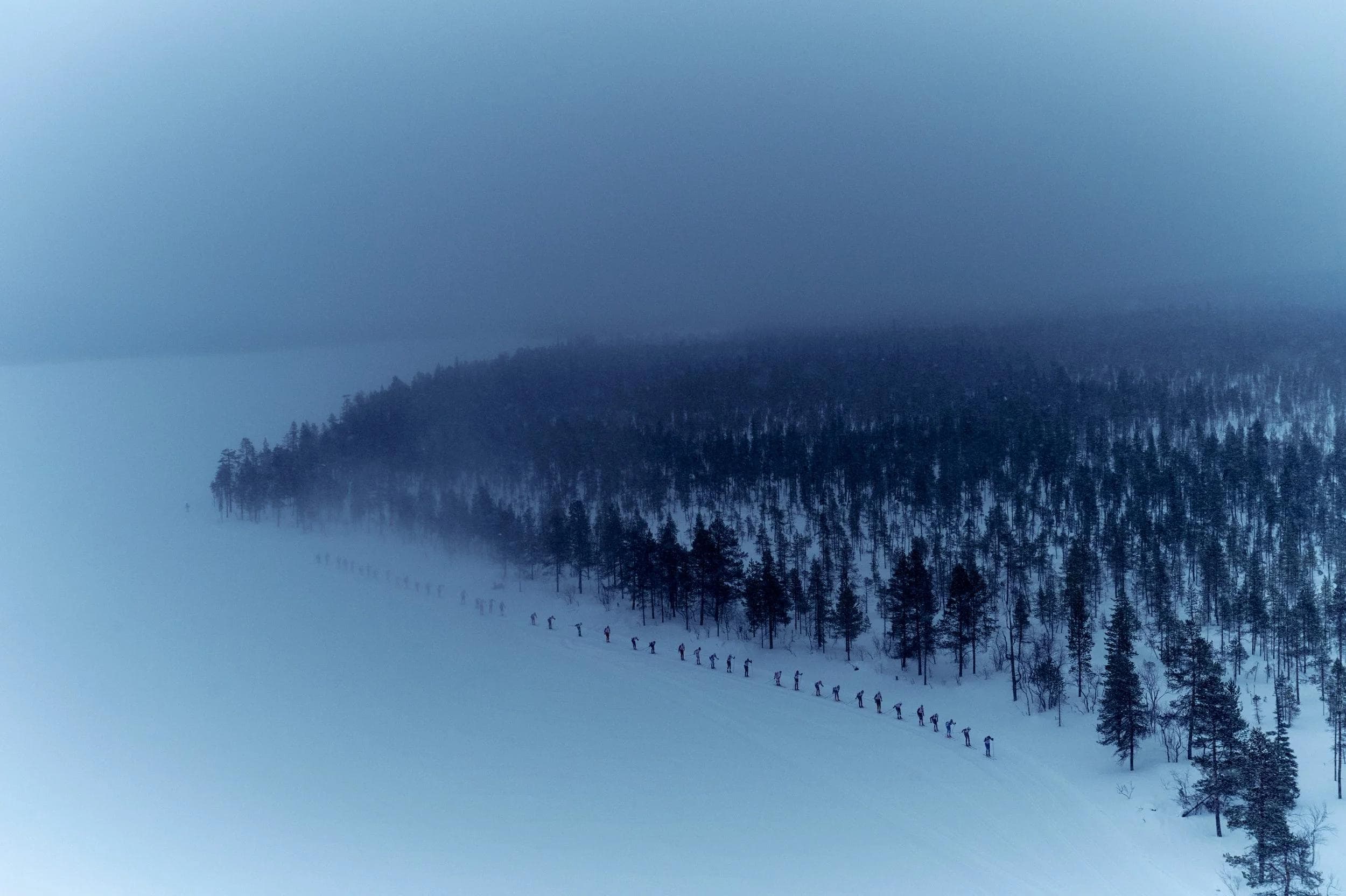 A long line of skiers moves across a snowy landscape beside a dark forest under a hazy sky.