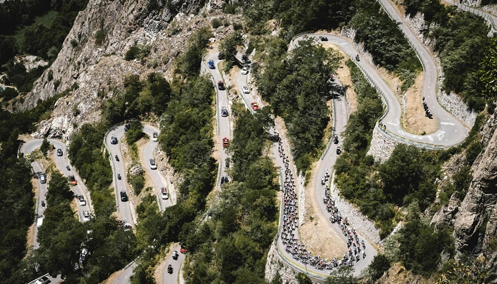 A cyclist in a yellow jersey climbs a mountain road, followed by a motorcycle, against a backdrop of steep valleys and jagged peaks.