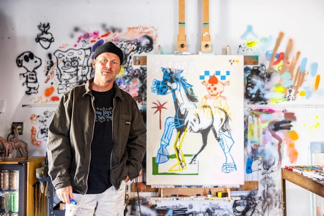 An artist in a beanie and jacket stands in his vibrant studio, featuring a large graphic painting of a horse and graffiti-covered walls.