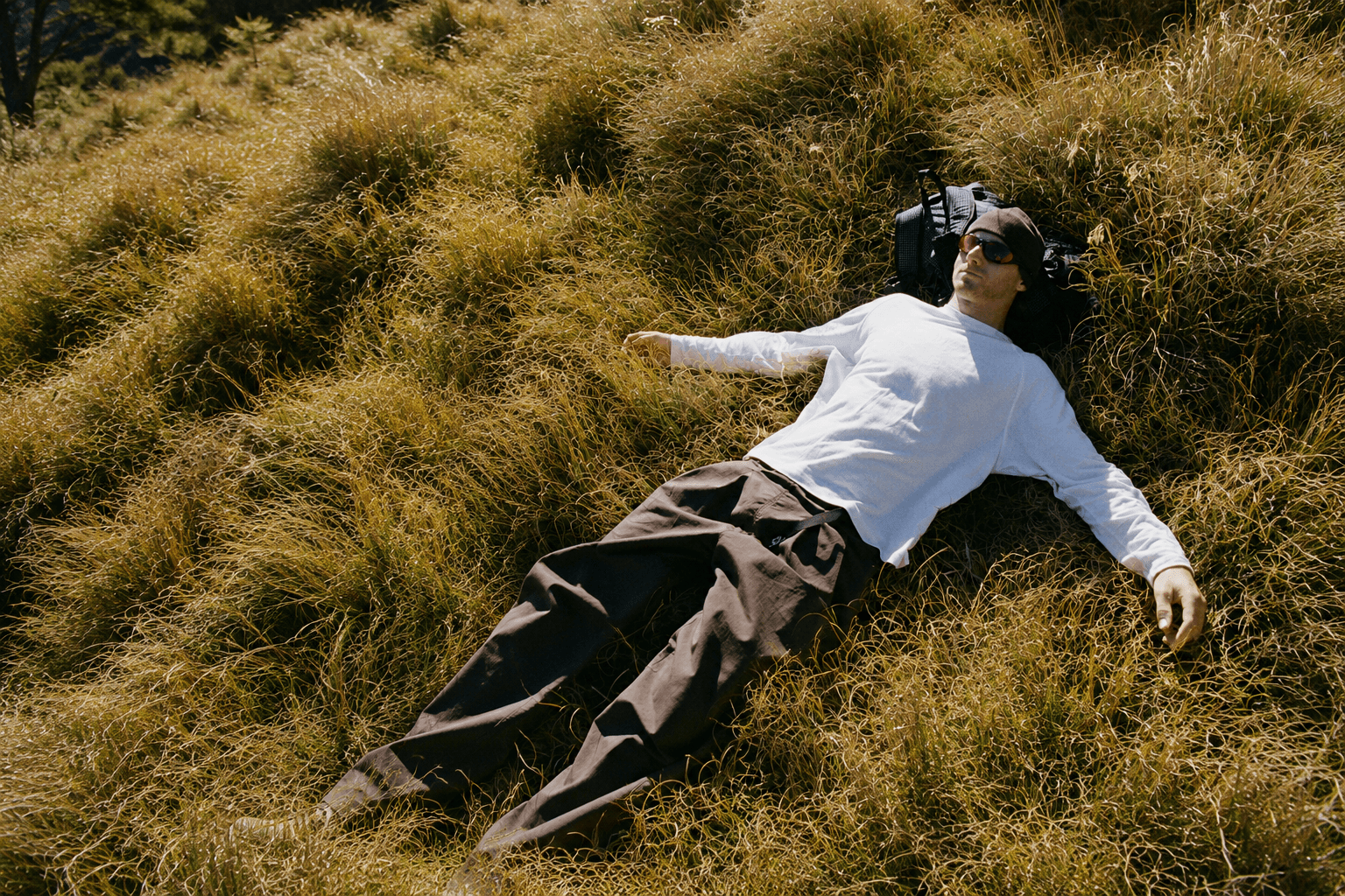 A man in a white shirt, beanie, and sunglasses rests on his back in dry, golden grass, using a backpack as a pillow.