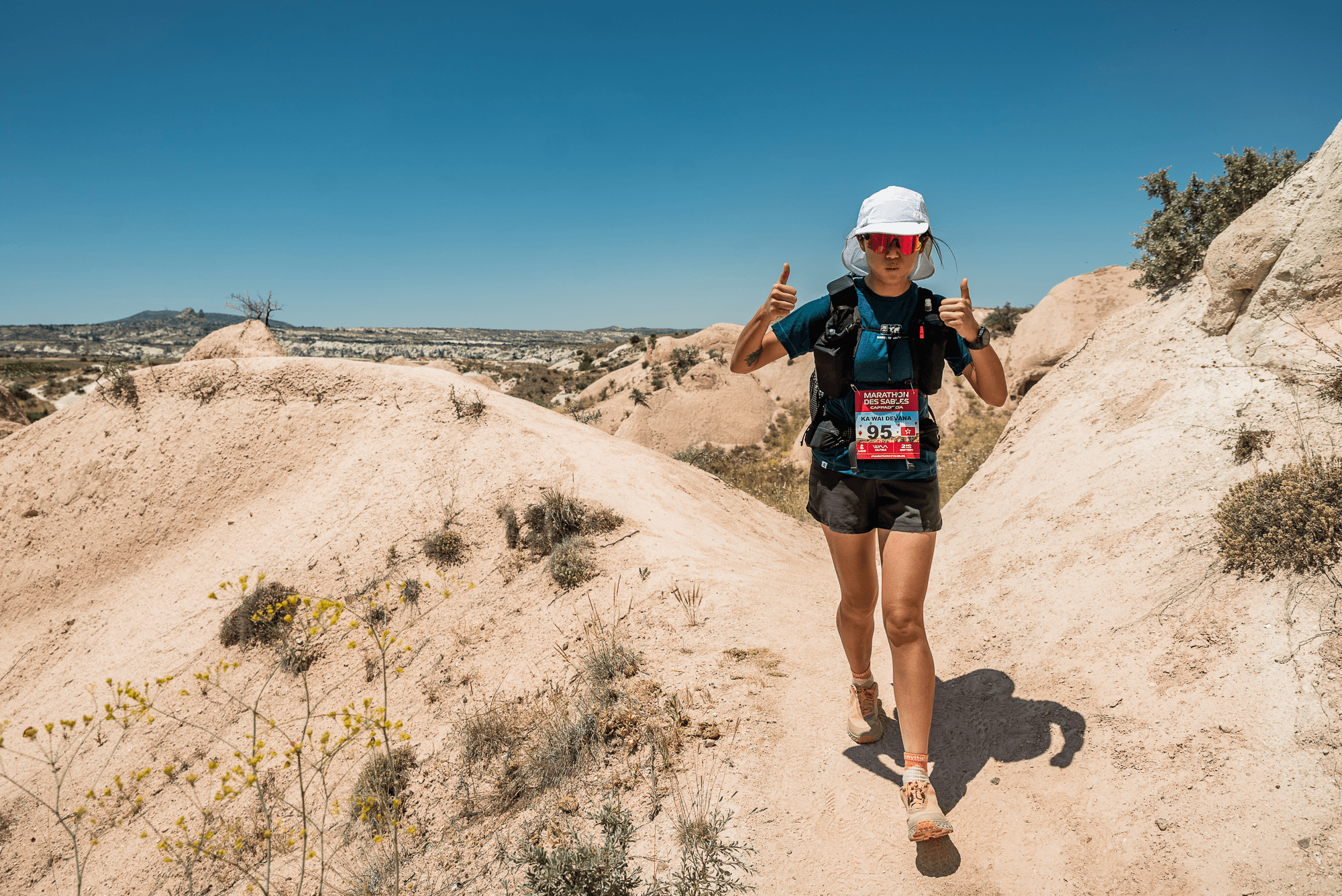 A trail runner in a race bib gives two thumbs up on a dusty desert path.