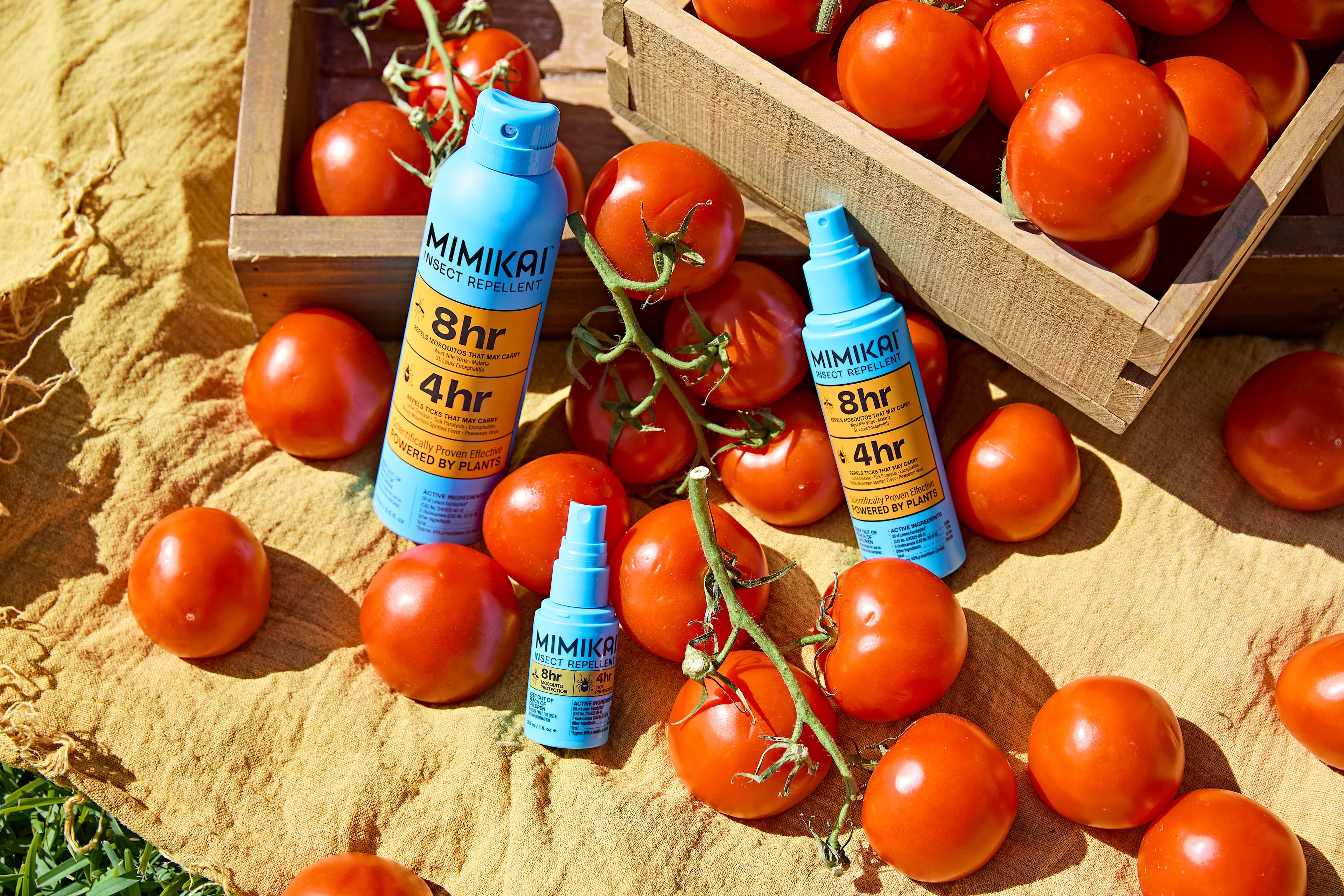 Three blue MIMIKAI insect repellent bottles in various sizes surrounded by red tomatoes on a yellow cloth.