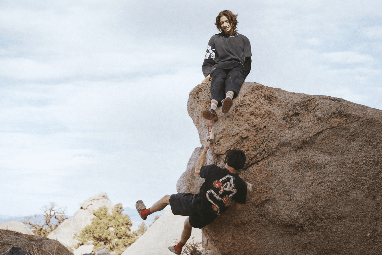 A person atop a boulder extends a hand to help another person climb up.