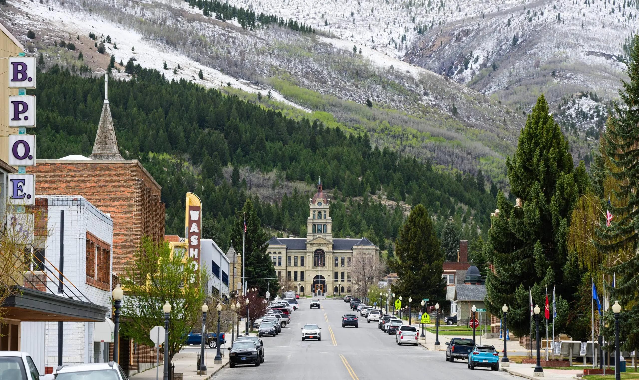 A street in a mountain town leading towards a large historic building, framed by snow-dusted, forested mountains.