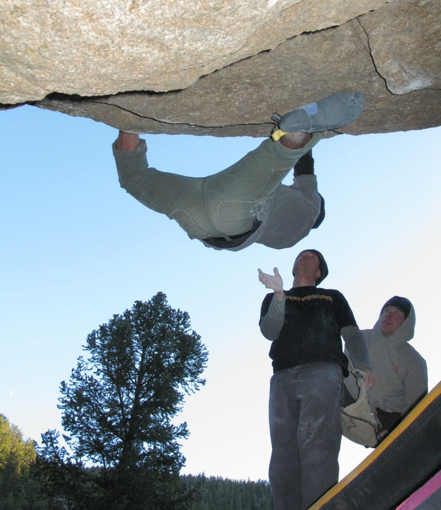 A boulderer hangs inverted from a rock overhang as two spotters look up.