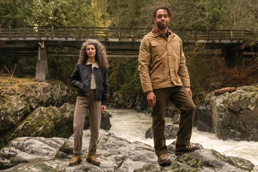 A man and a woman stand on rocks beside a rushing river, with a wooden bridge in the background.