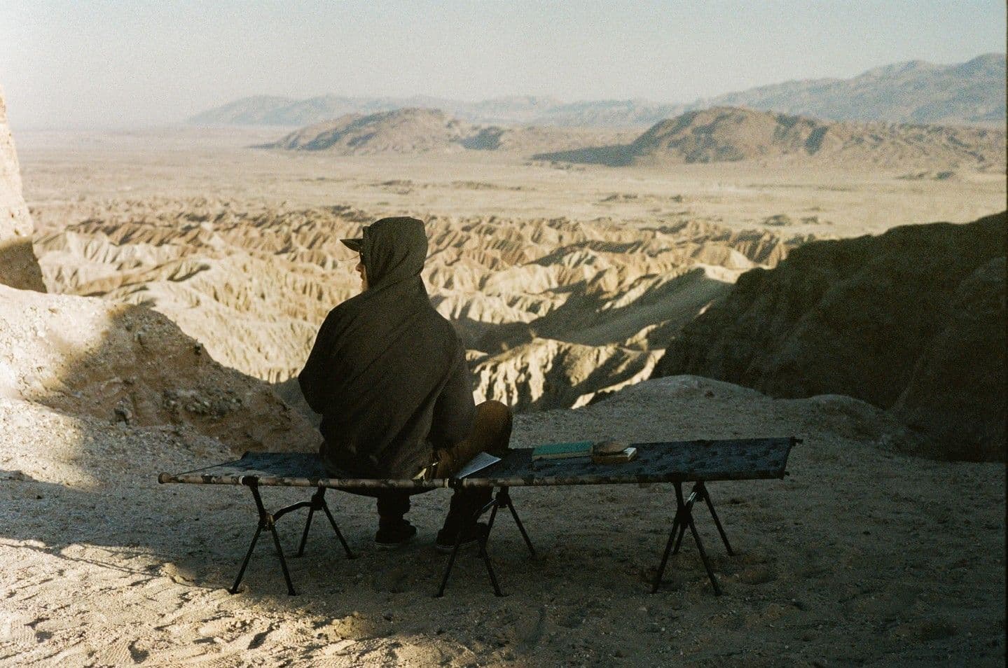 A person in a hoodie sits on a camping cot, gazing at a vast, rugged desert landscape.