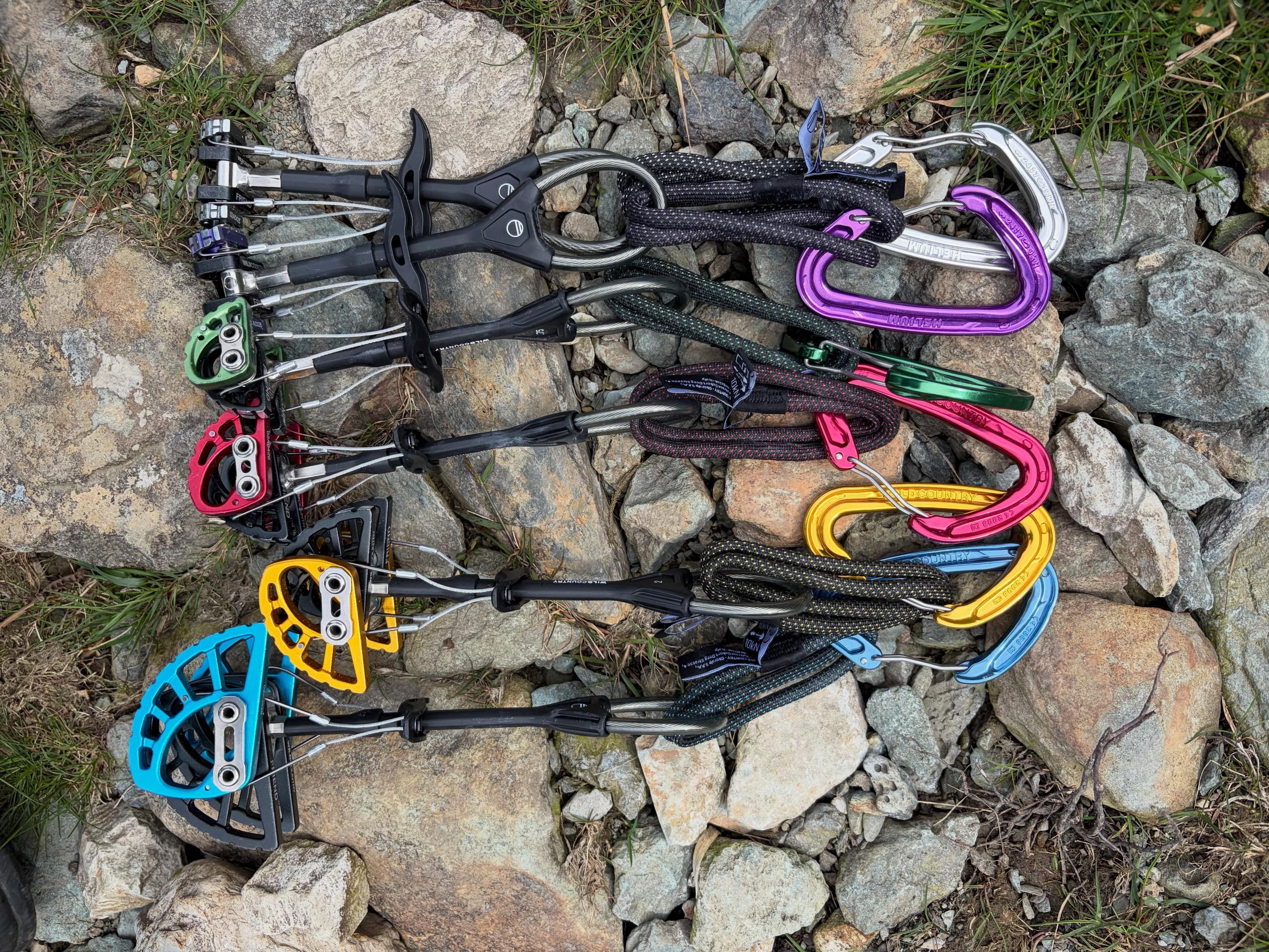 A set of five colorful climbing cams with attached slings and carabiners, laid on rocks.