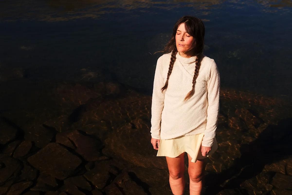 Woman with two braids stands in shallow water, eyes closed.